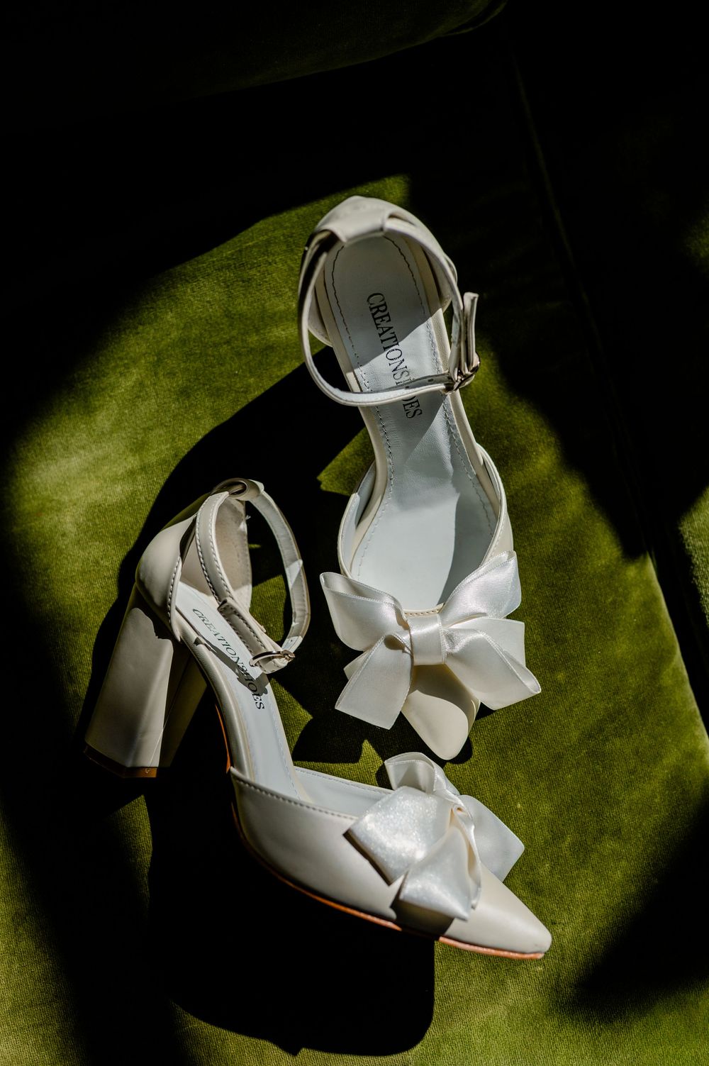 White satin block heel wedding shoes with bow detail and ankle strap, displayed on green velvet surface.