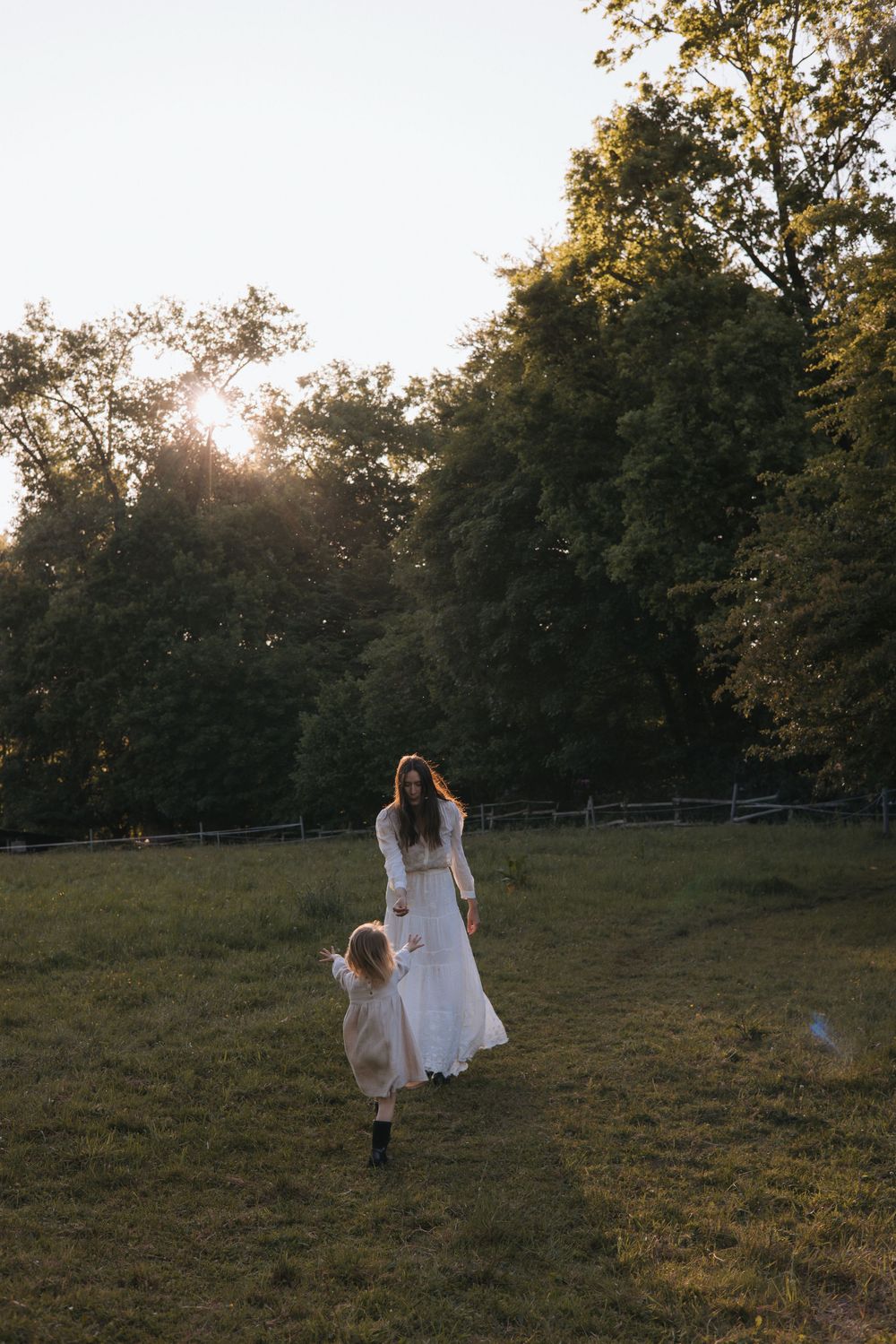 A mother in a white dress holds her young daughter's hand while walking through a sunlit green meadow at golden hour.