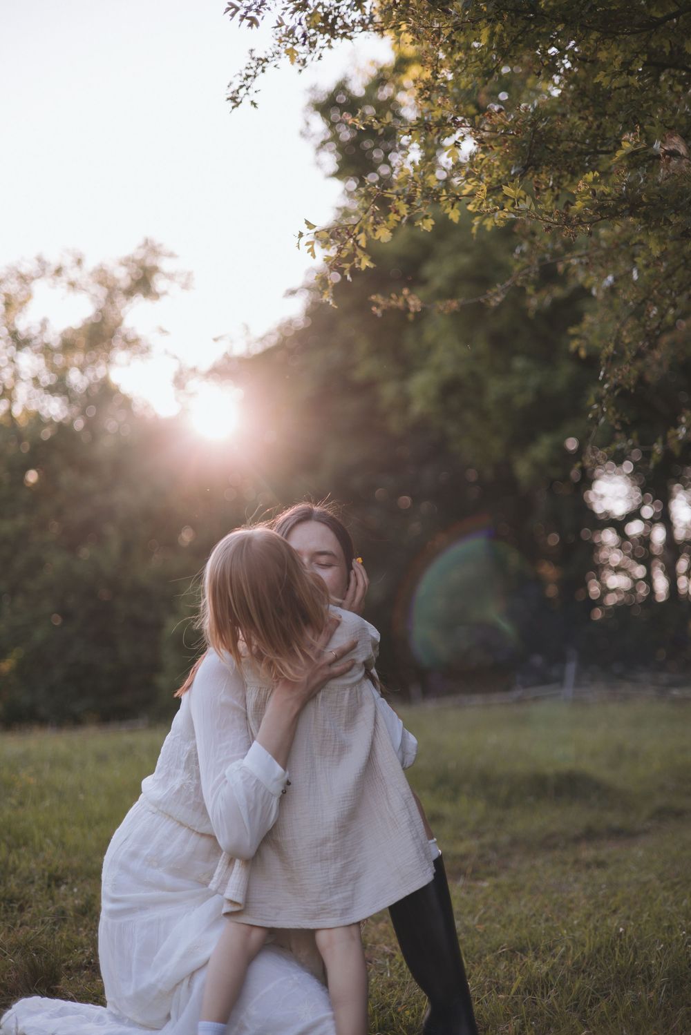 Two women embracing tenderly in a sunlit meadow with golden hour light filtering through green trees.