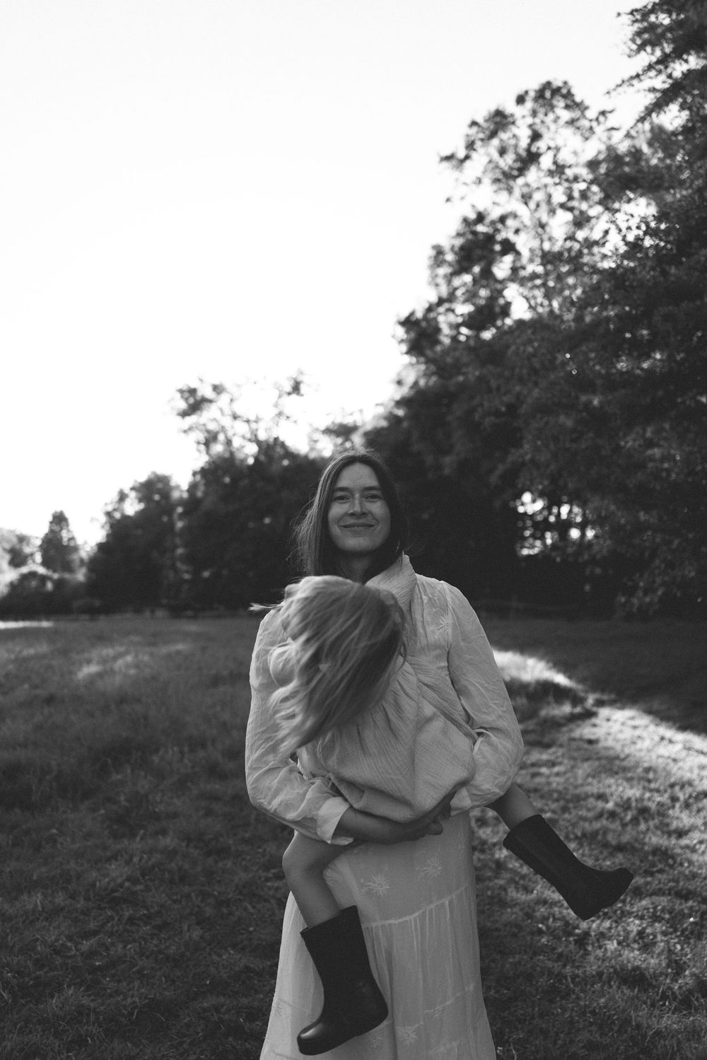 Black and white photo of a smiling woman holding a child outdoors near trees on a grassy path.