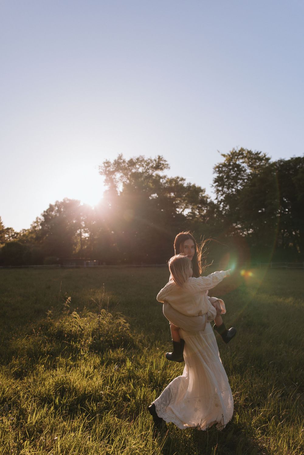 Woman in white dress carrying a child in a sunlit green field with trees in the background.