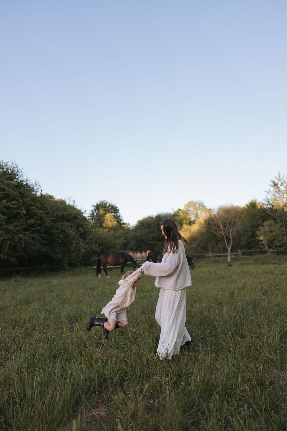 Woman in white dress spinning young child in green meadow with horse and trees in background at dusk.