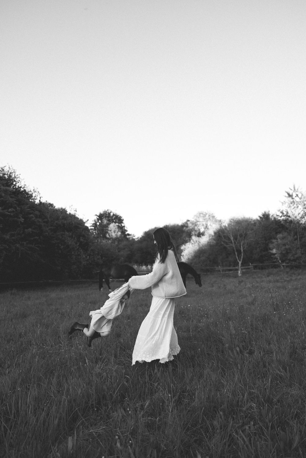 Black and white photo of a woman in a white dress twirling a child in a grassy field with trees in the background.