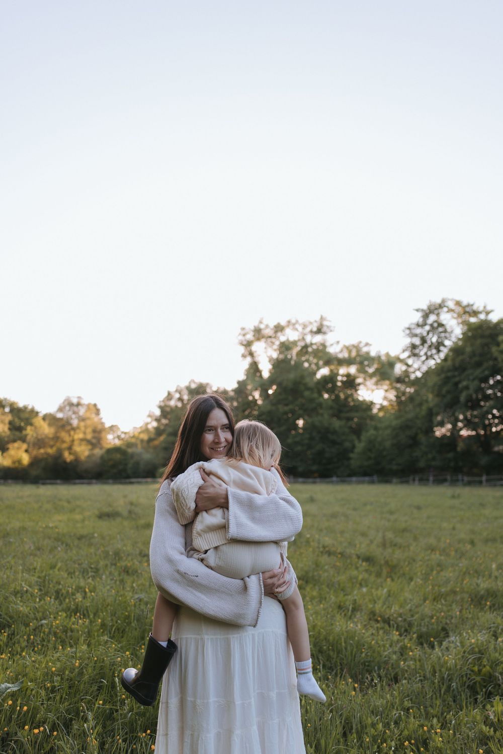 A smiling mother in a white dress lovingly holds her toddler in a lush green field during sunset.