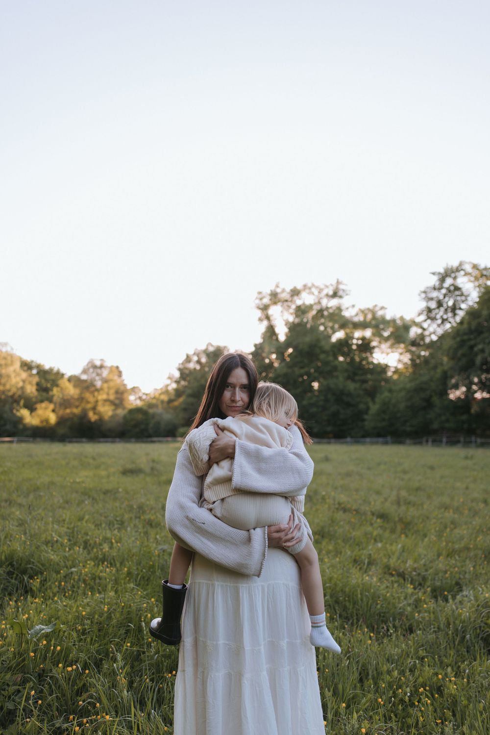 A woman in a white dress holds a toddler in a green field at dusk, with trees in the background.