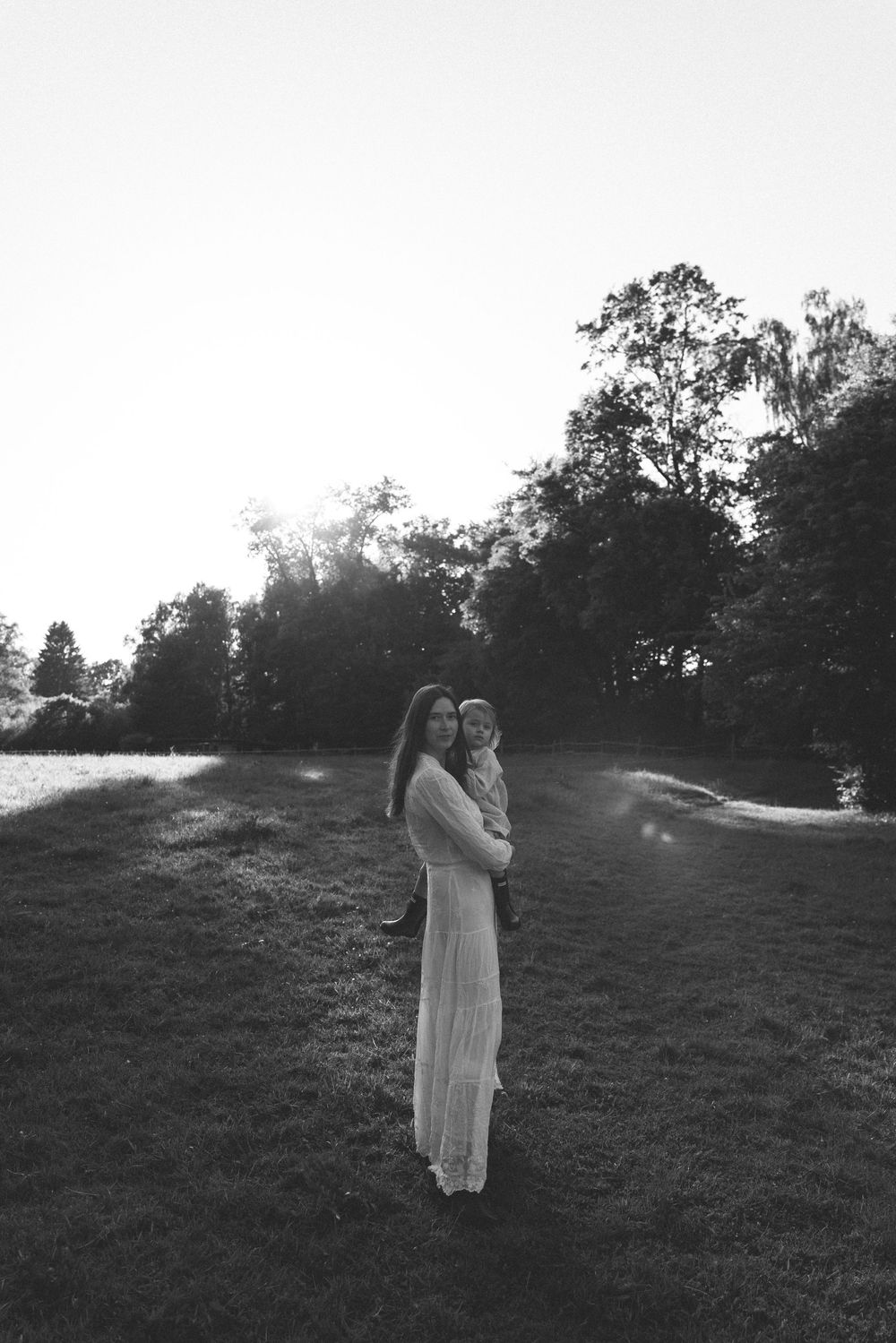 Black and white photo of a mother in a long dress holding her child in a sunlit park with lush trees.