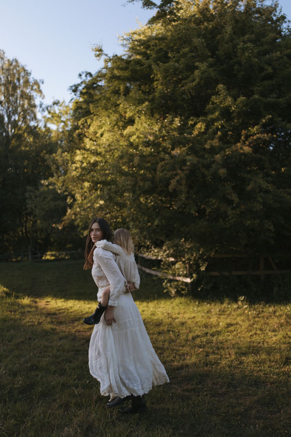 Woman in white lace dress holding a child in a sunlit green park.