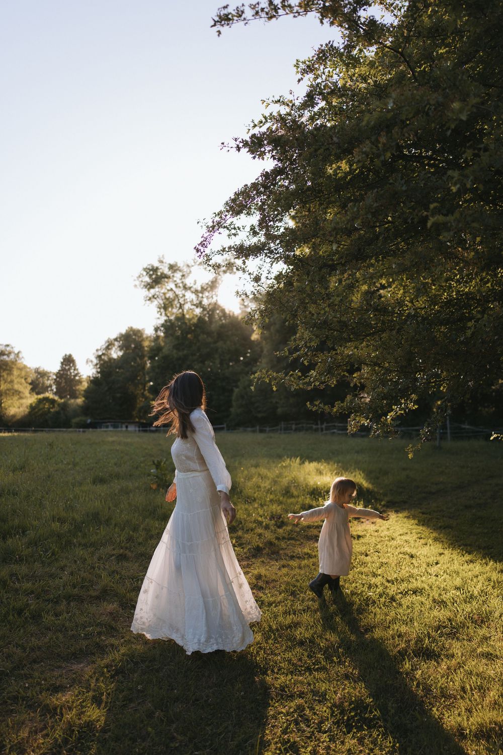 Bride in white lace gown walks with flower girl across sunlit green meadow surrounded by tall trees at golden hour.