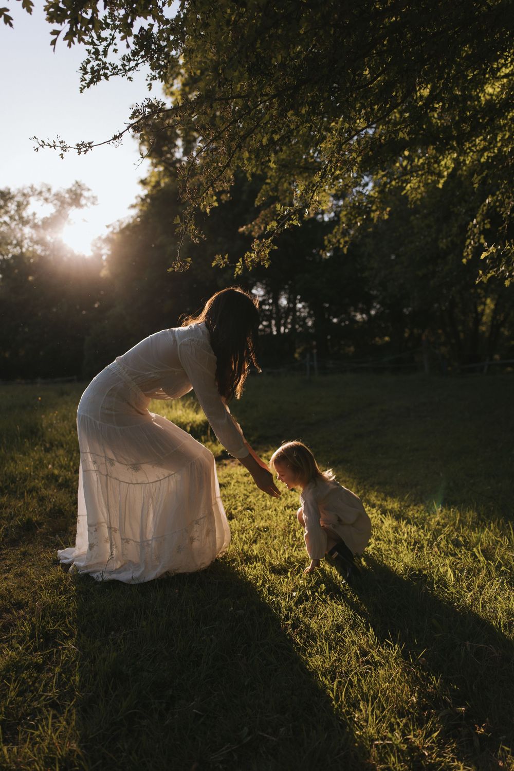 Mother in white dress helping toddler walk on green grass during golden hour sunset in a park.