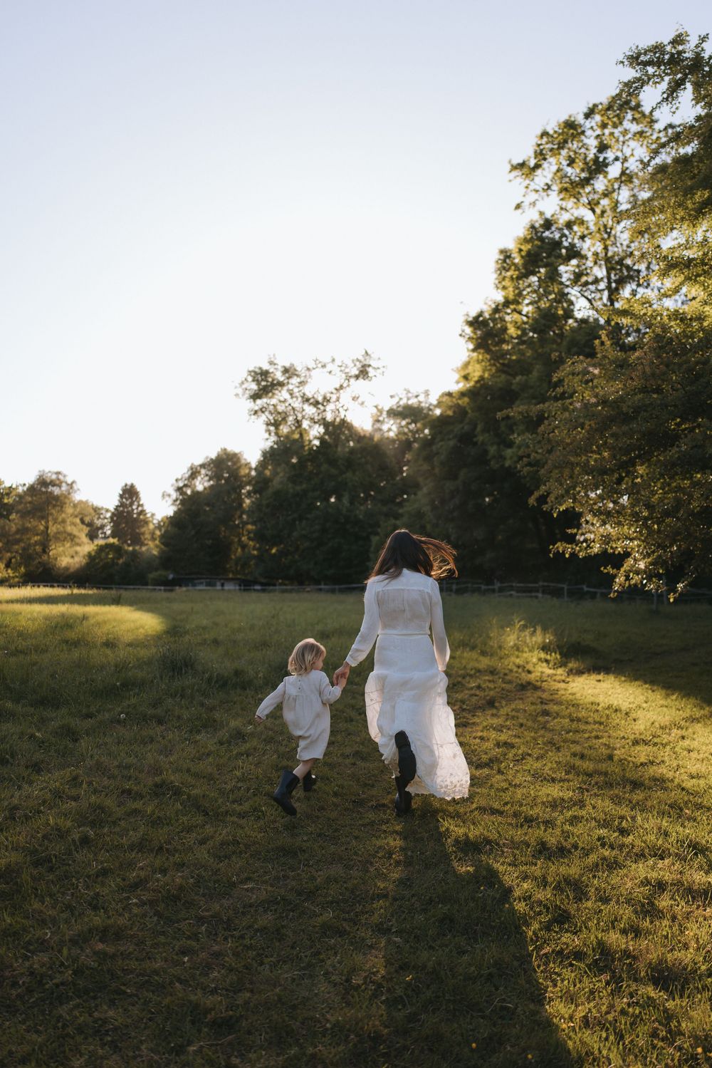 A bride in a white gown holds hands with a young child running through a sunlit green meadow surrounded by trees.