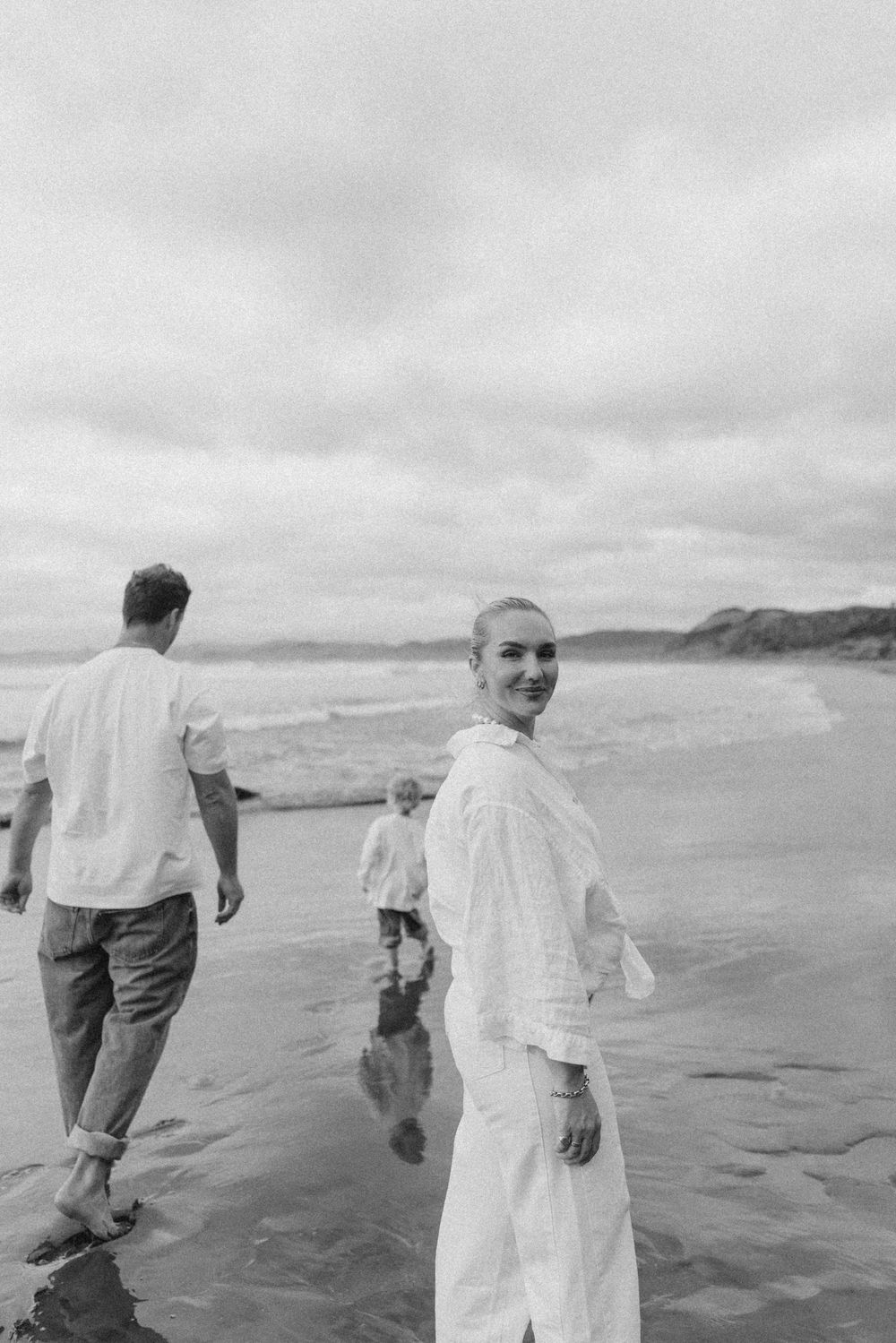 Woman in white dress smiles on a beach in black and white photo with others walking near shoreline.