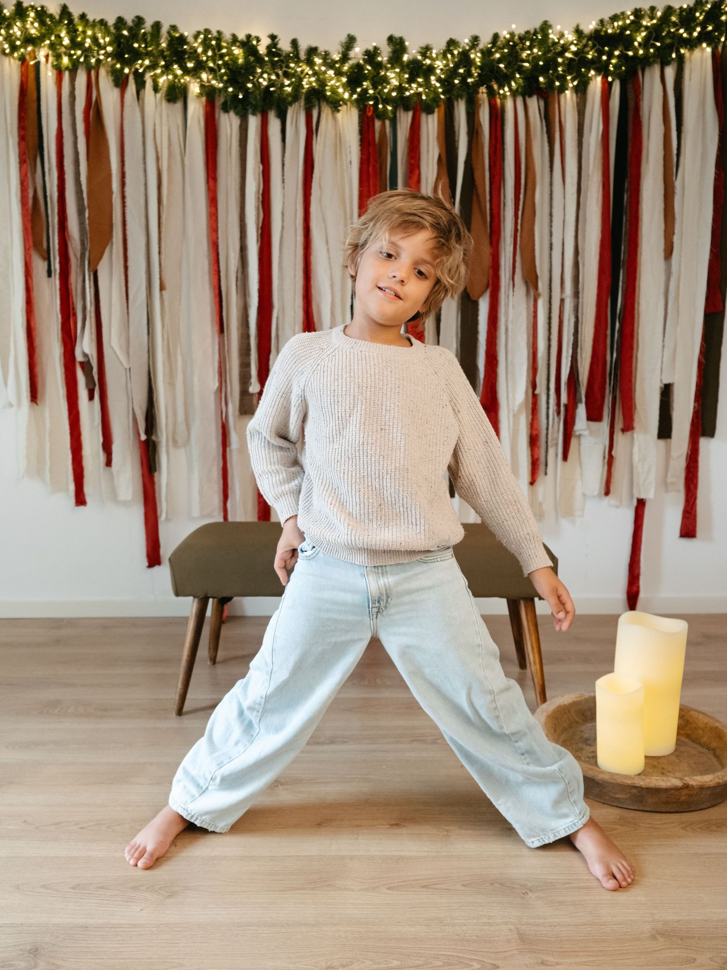 A child in a beige sweater and light jeans poses confidently in front of a festive Christmas ribbon backdrop.