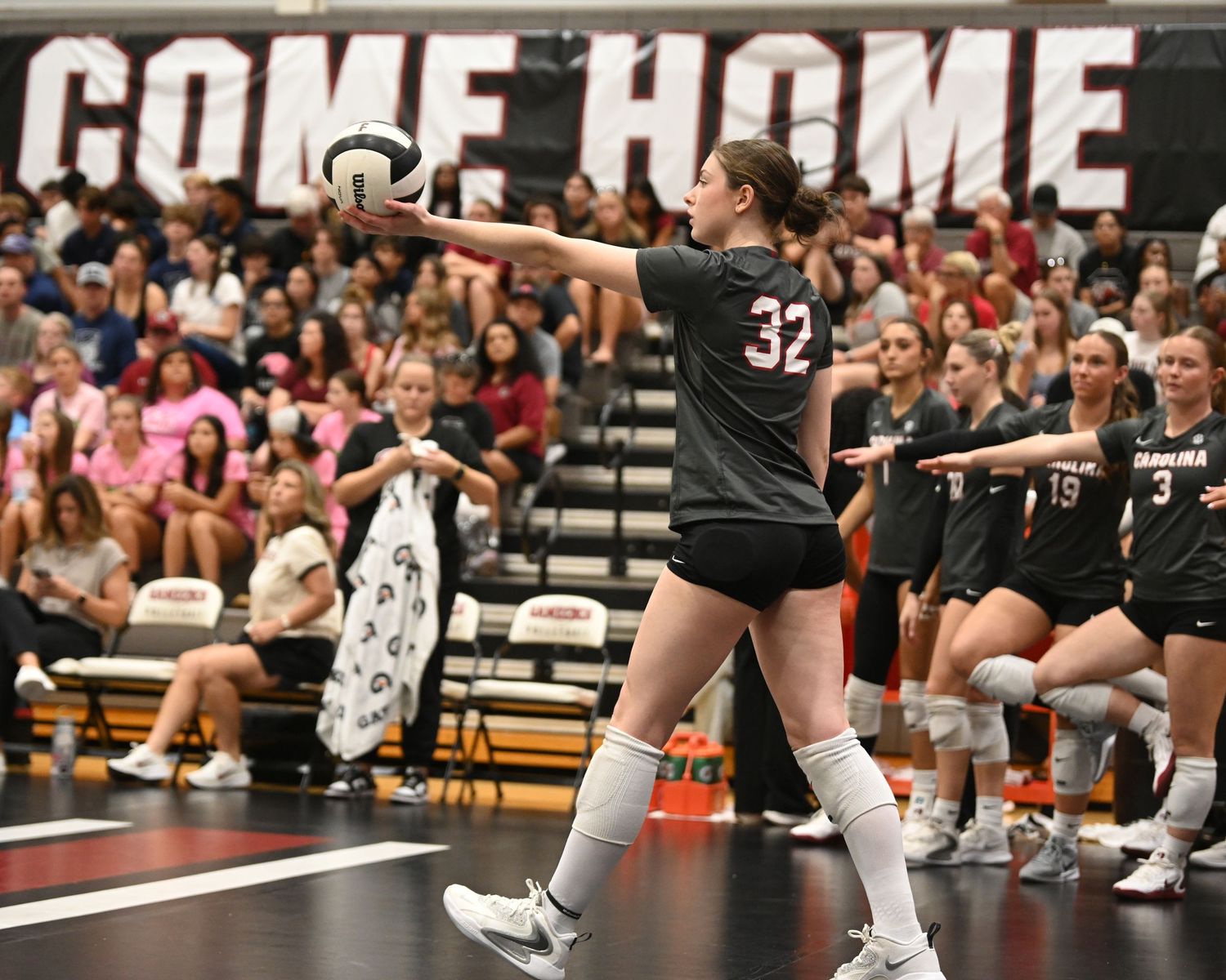 Volleyball player wearing number 32 points toward the court in front of her teammates and a home-crowd banner.
