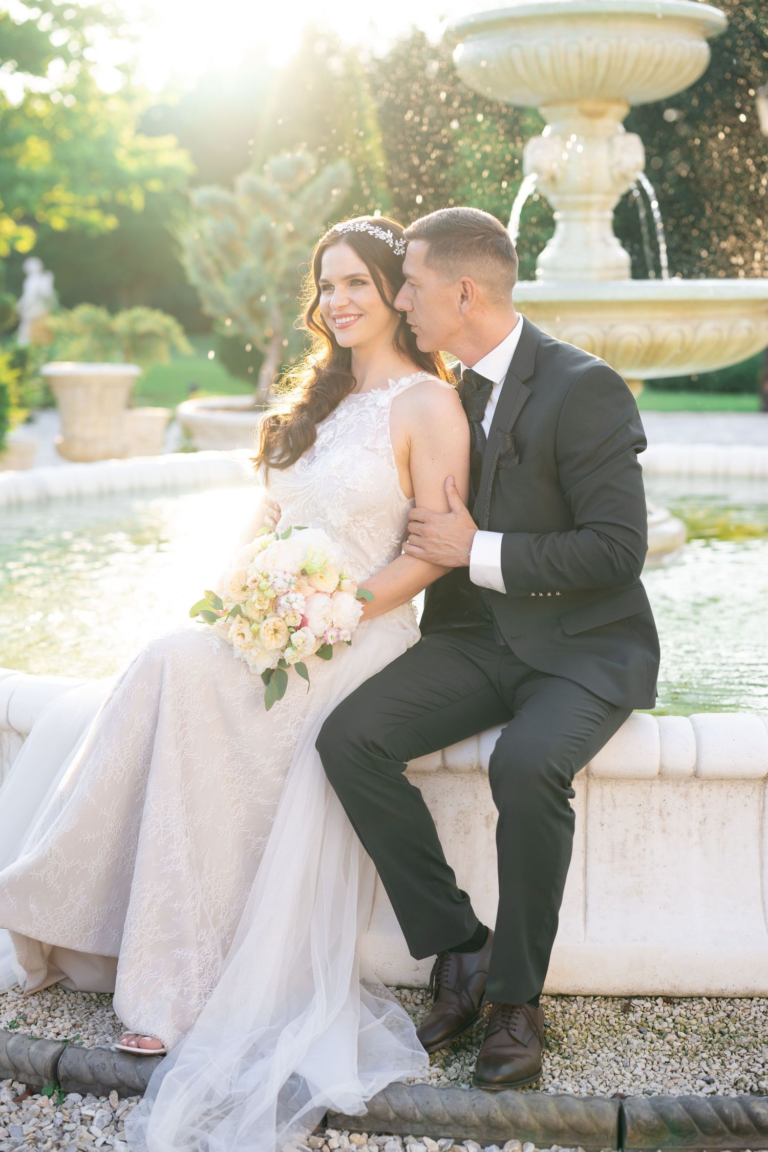Bride and groom sit by ornate stone fountain in golden sunlight, bride holding pink bouquet.