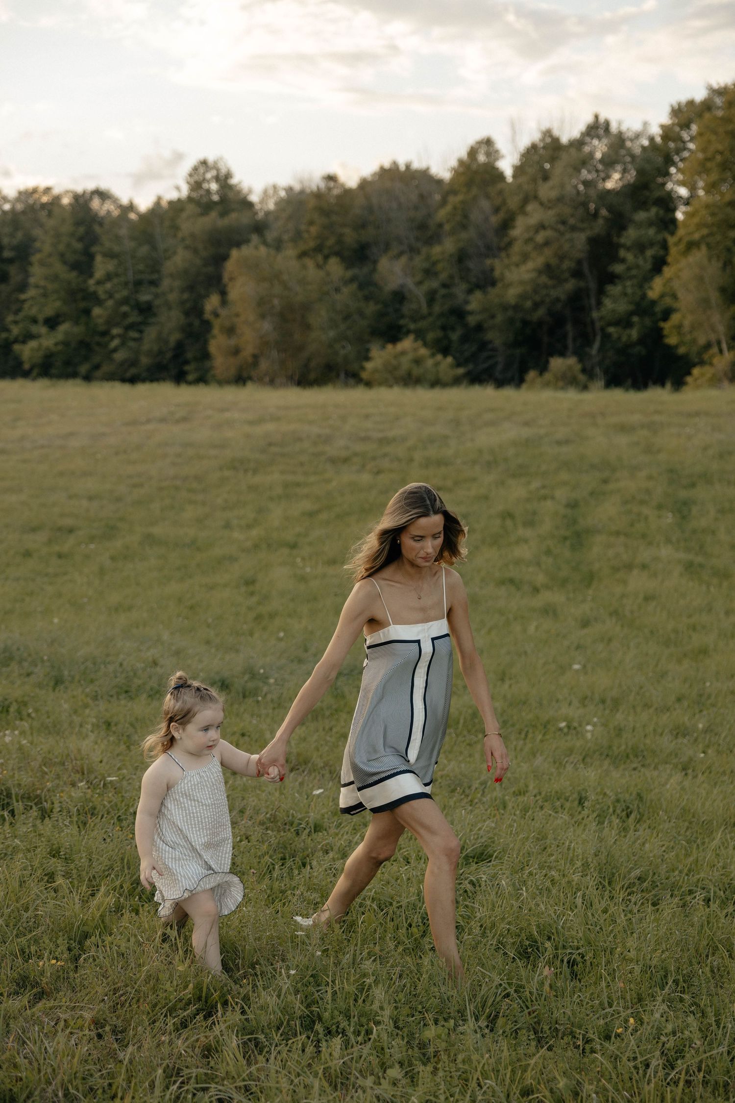 A woman and young girl hold hands walking through a lush green field with trees in the background.