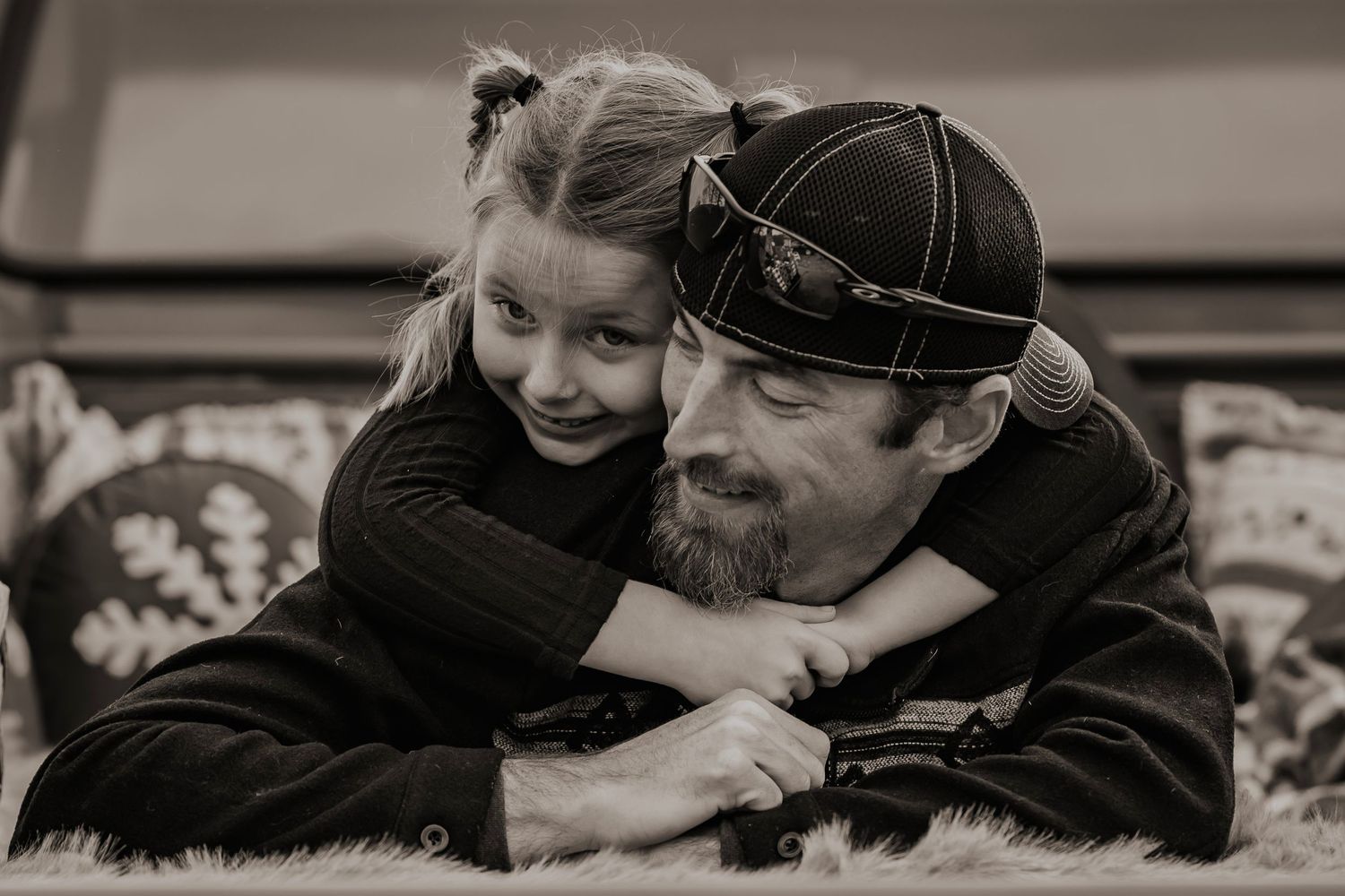 Black and white photo of a bearded man in a cap smiling as a young girl hugs him from behind outdoors.