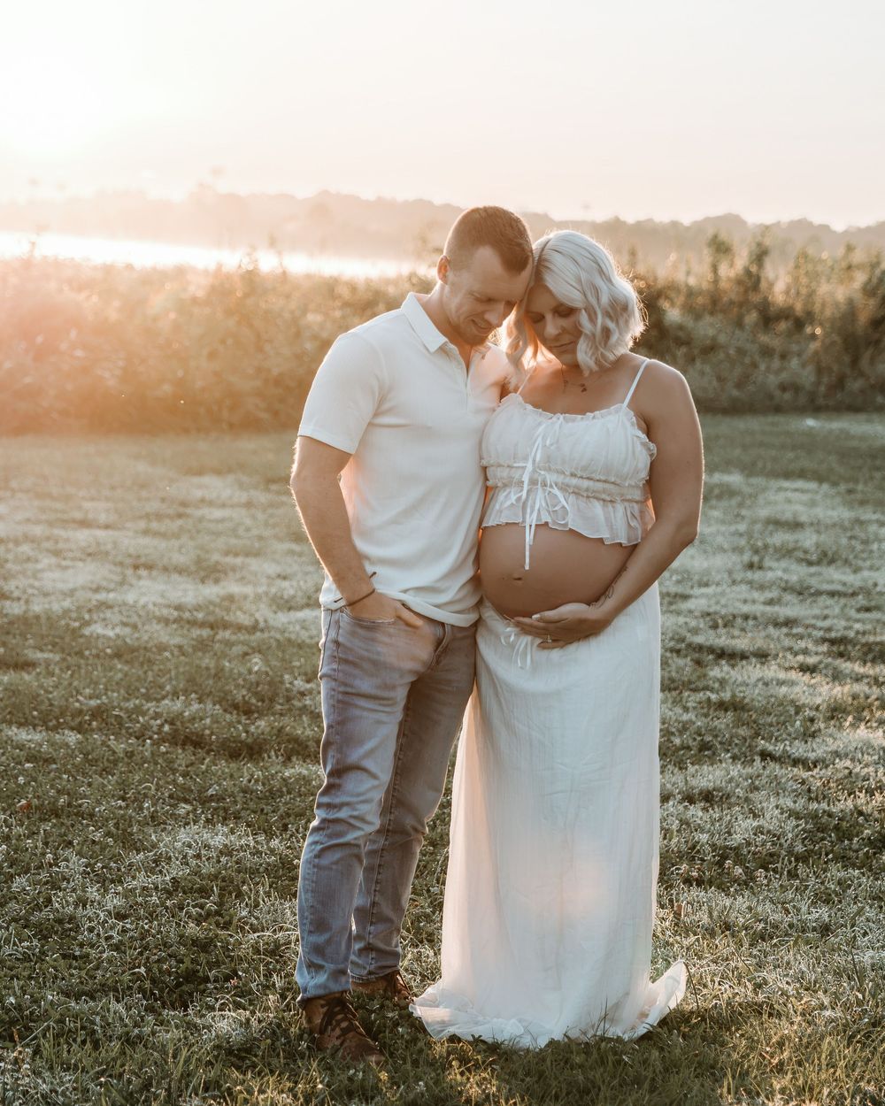 Couple poses for golden hour maternity photo shoot outdoors, woman in white dress cradling baby bump.