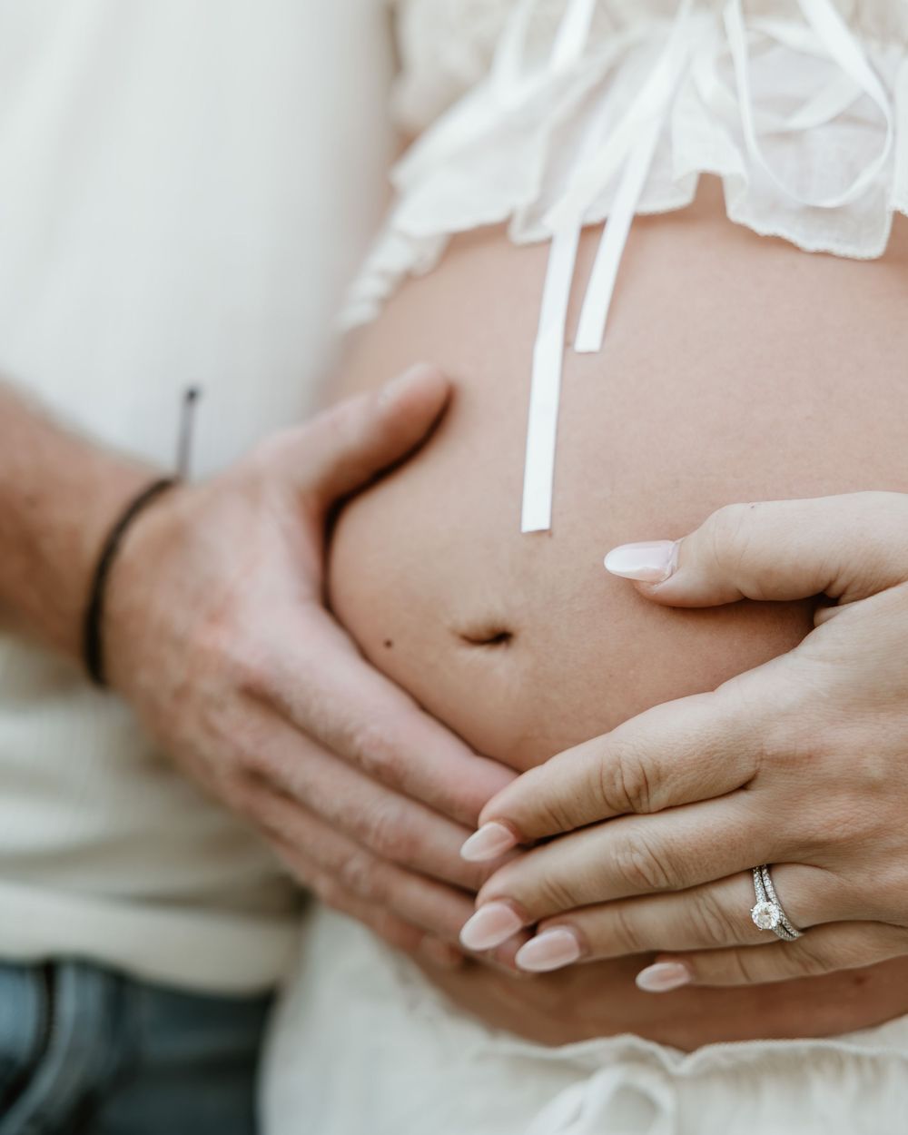 Couple tenderly cradling pregnant belly, woman wearing engagement ring, soft white maternity dress, close-up pregnancy photo.