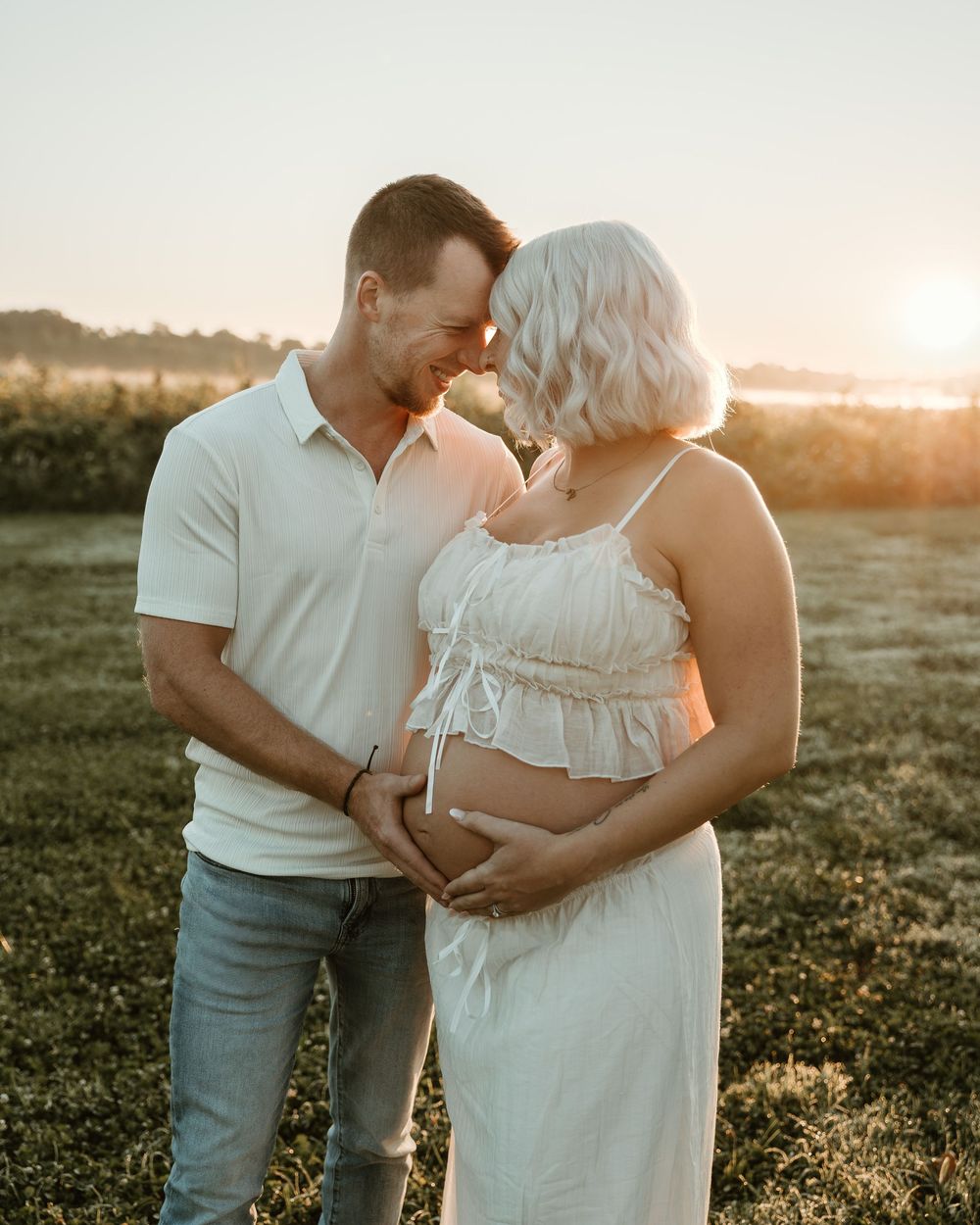 Couple cradling pregnant belly at golden hour in a sunlit field, wearing white outfits, sharing a tender forehead-to-forehead moment.
