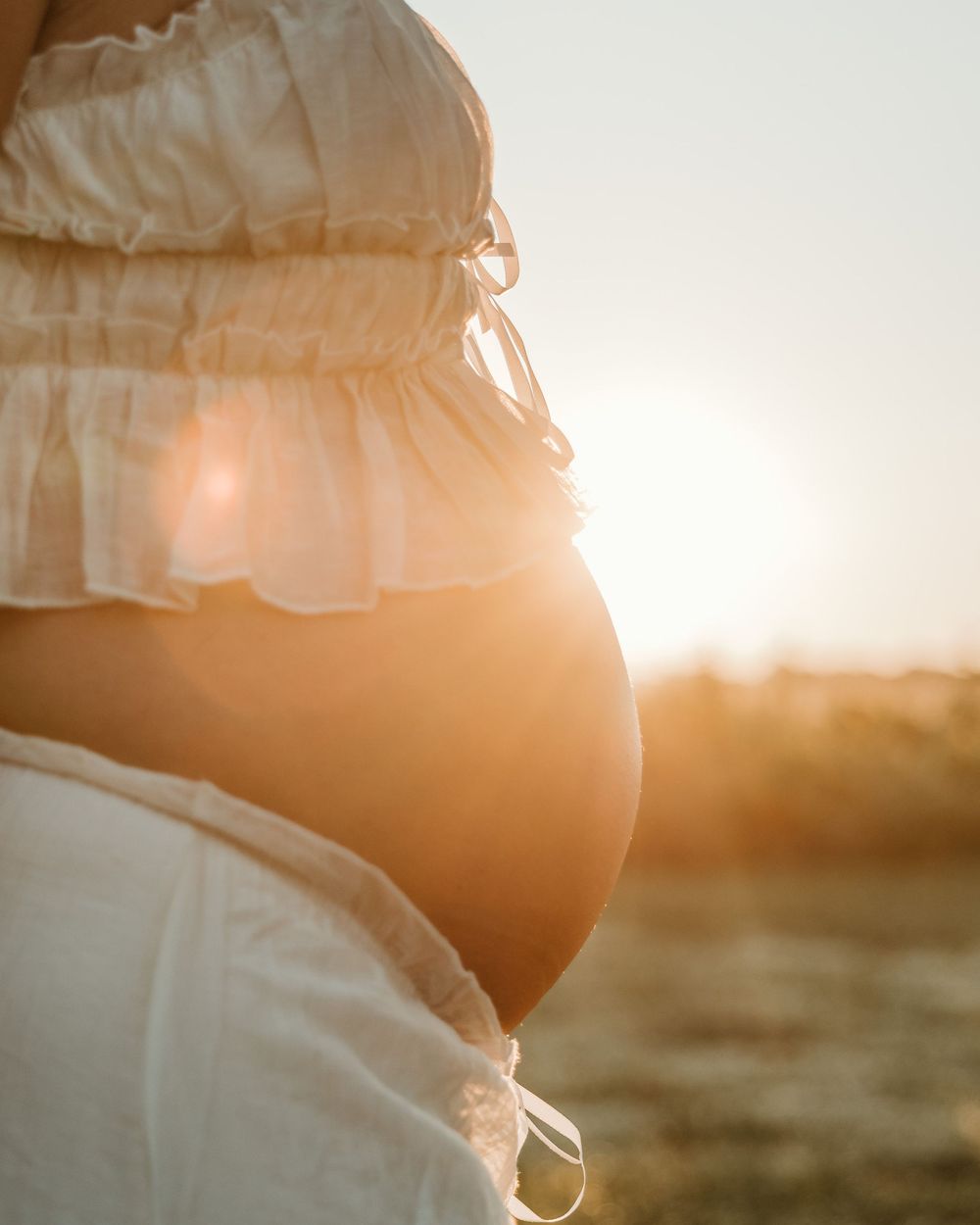 Pregnant woman in white ruffled crop top with bare belly glowing in golden sunset light outdoors.