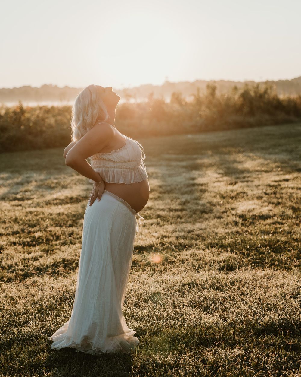 Pregnant woman in white dress standing in sunlit field at golden hour, cradling baby bump during maternity photoshoot.
