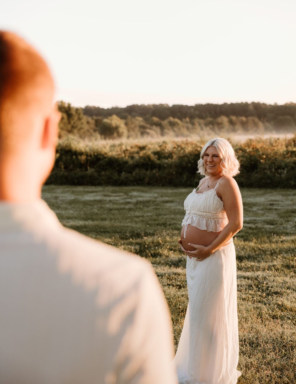 Pregnant woman in white maternity gown cradling baby bump in golden hour field with misty trees backdrop.