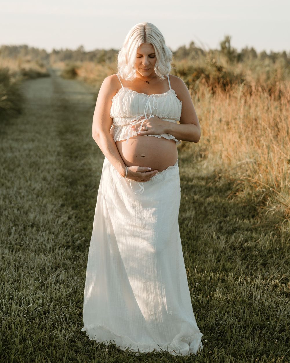 Pregnant woman in white maxi dress holding baby bump in golden hour field maternity photoshoot.