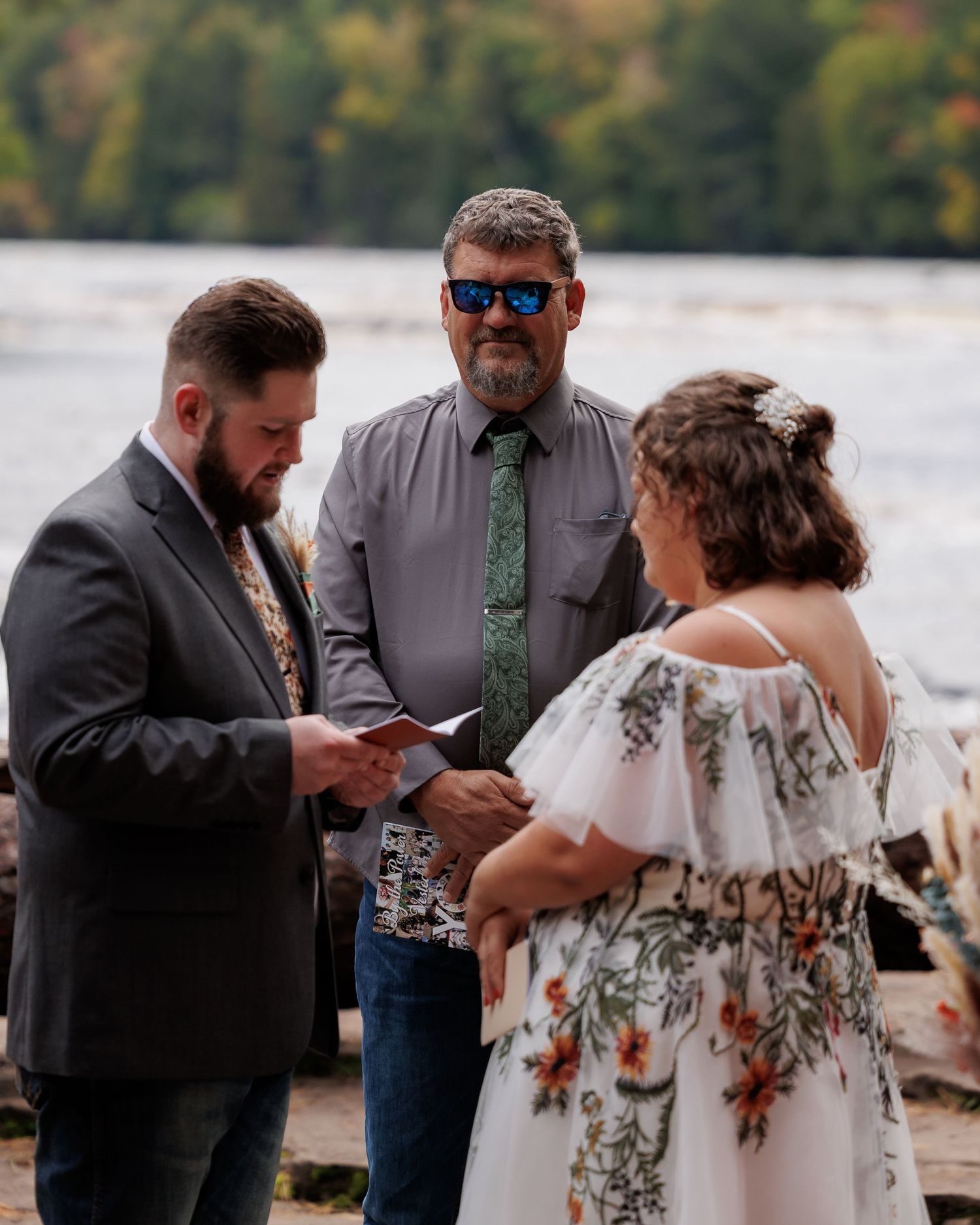 Outdoor wedding ceremony with bride in floral gown reading vows, officiant, groom, and bridesmaid in rust dress by a river.