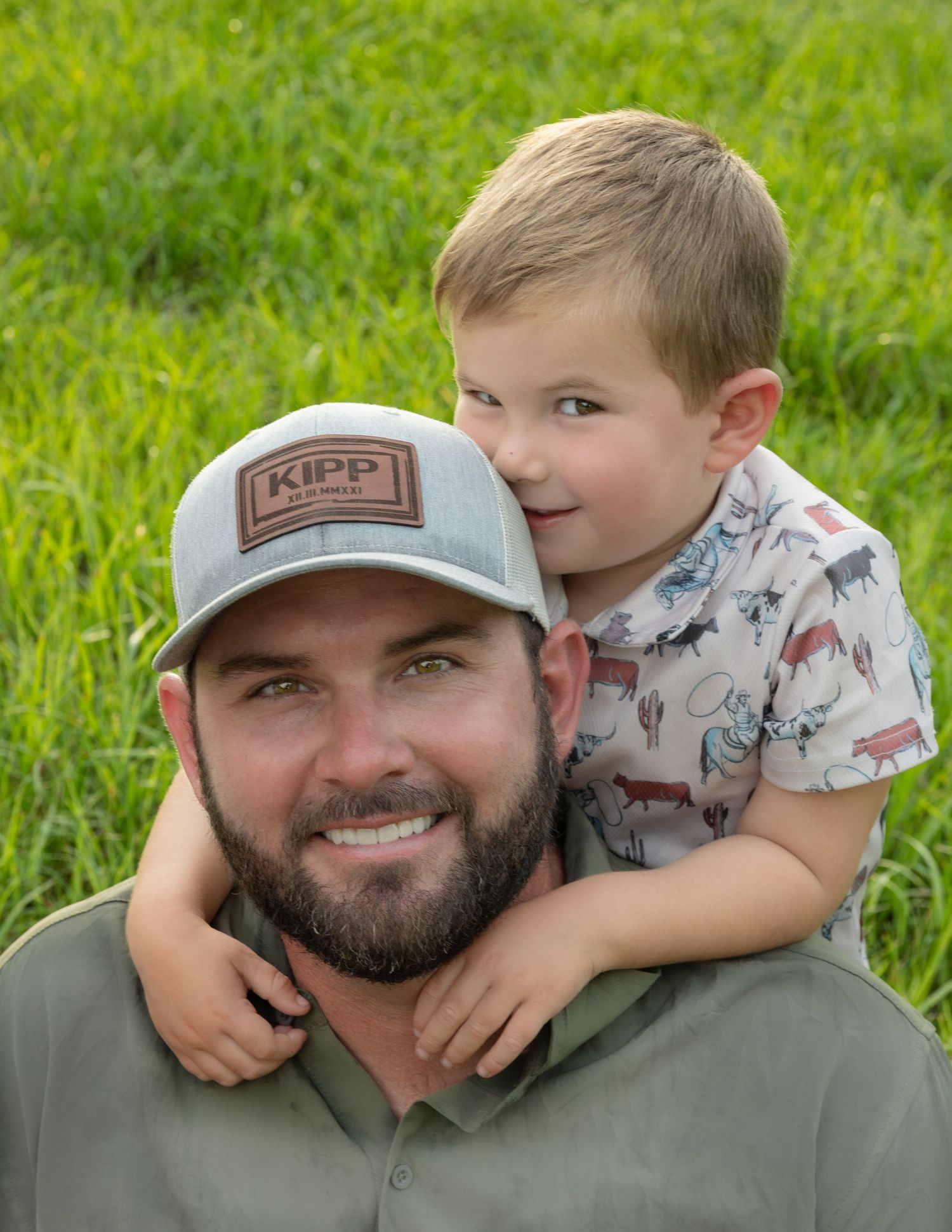 Smiling bearded man in a gray KIPP cap with a toddler boy hugging him from behind on his shoulders in a green grassy field.
