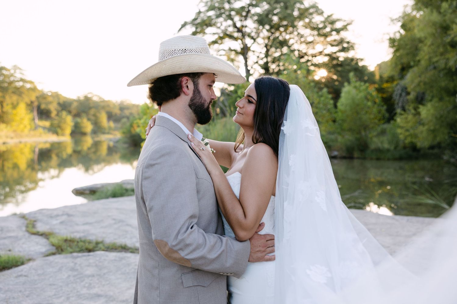 Bride and groom share a romantic moment outdoors near a river, groom wearing a cowboy hat and gray suit.