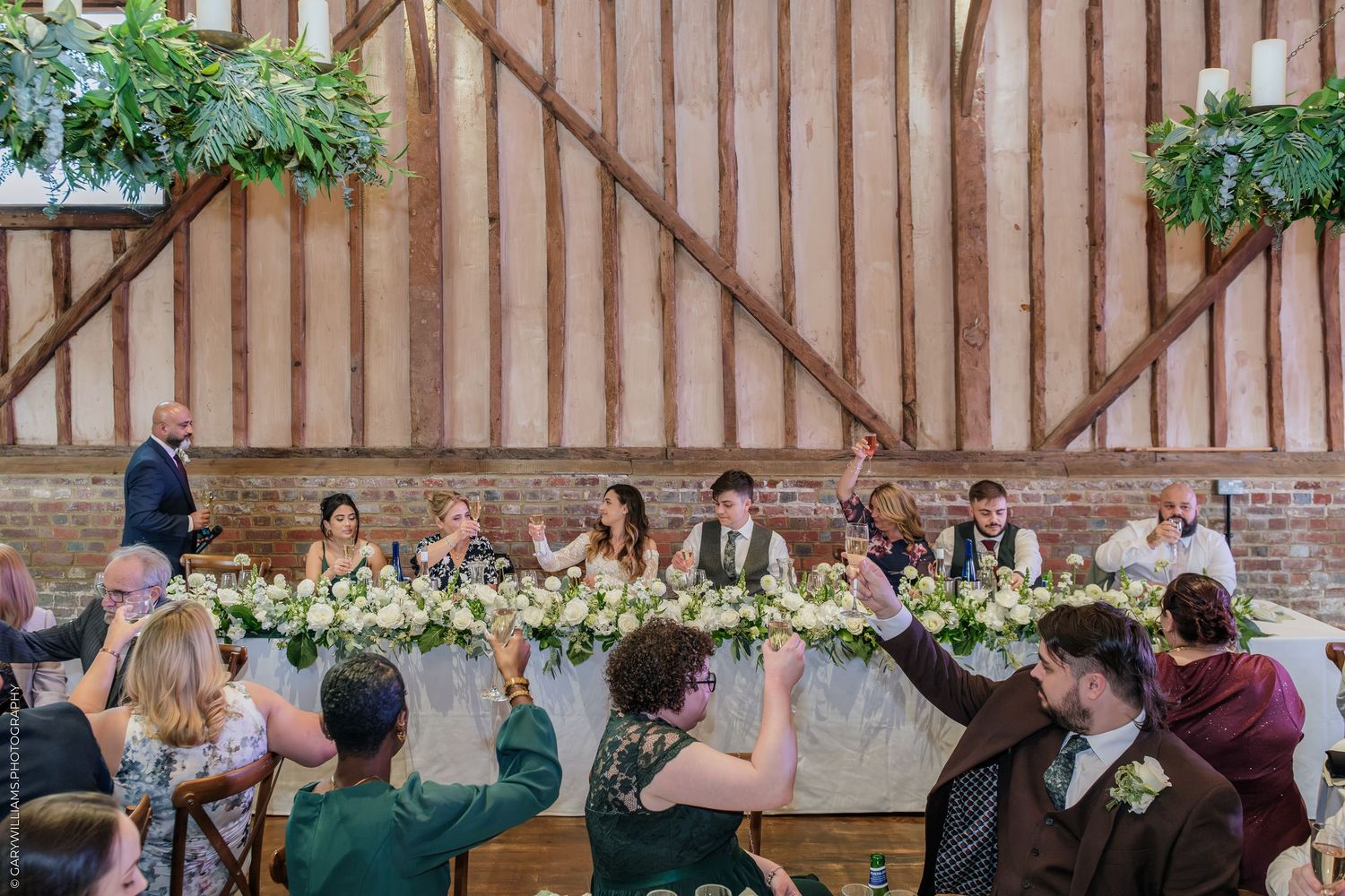 Wedding party toasts at a rustic barn reception, featuring a floral-adorned head table with greenery decor.