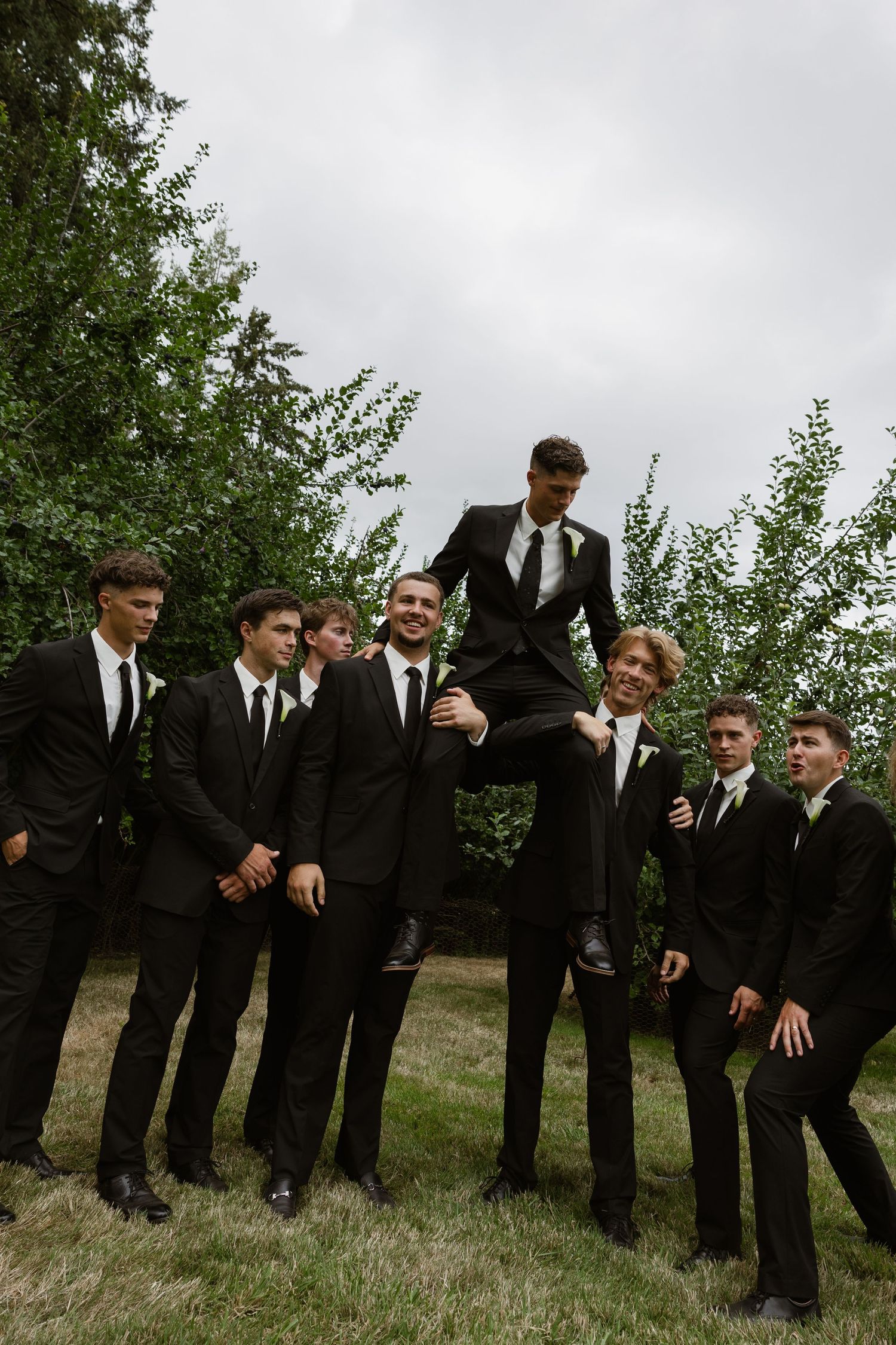 Groomsmen in black suits lift the groom outdoors, surrounded by lush green trees on a cloudy wedding day.
