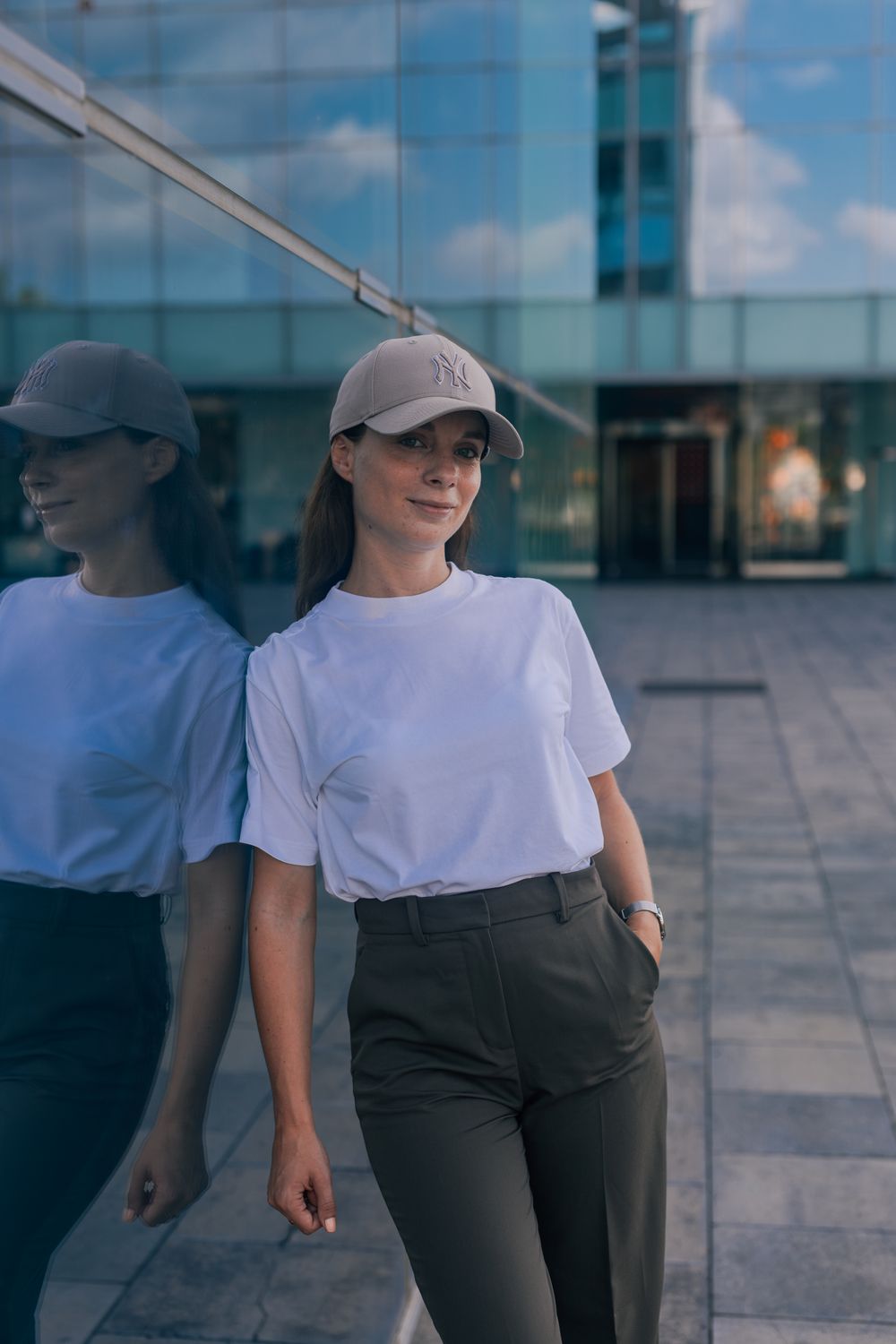 Young woman in white tee, khaki cap, and dark trousers posing against a reflective glass building facade.
