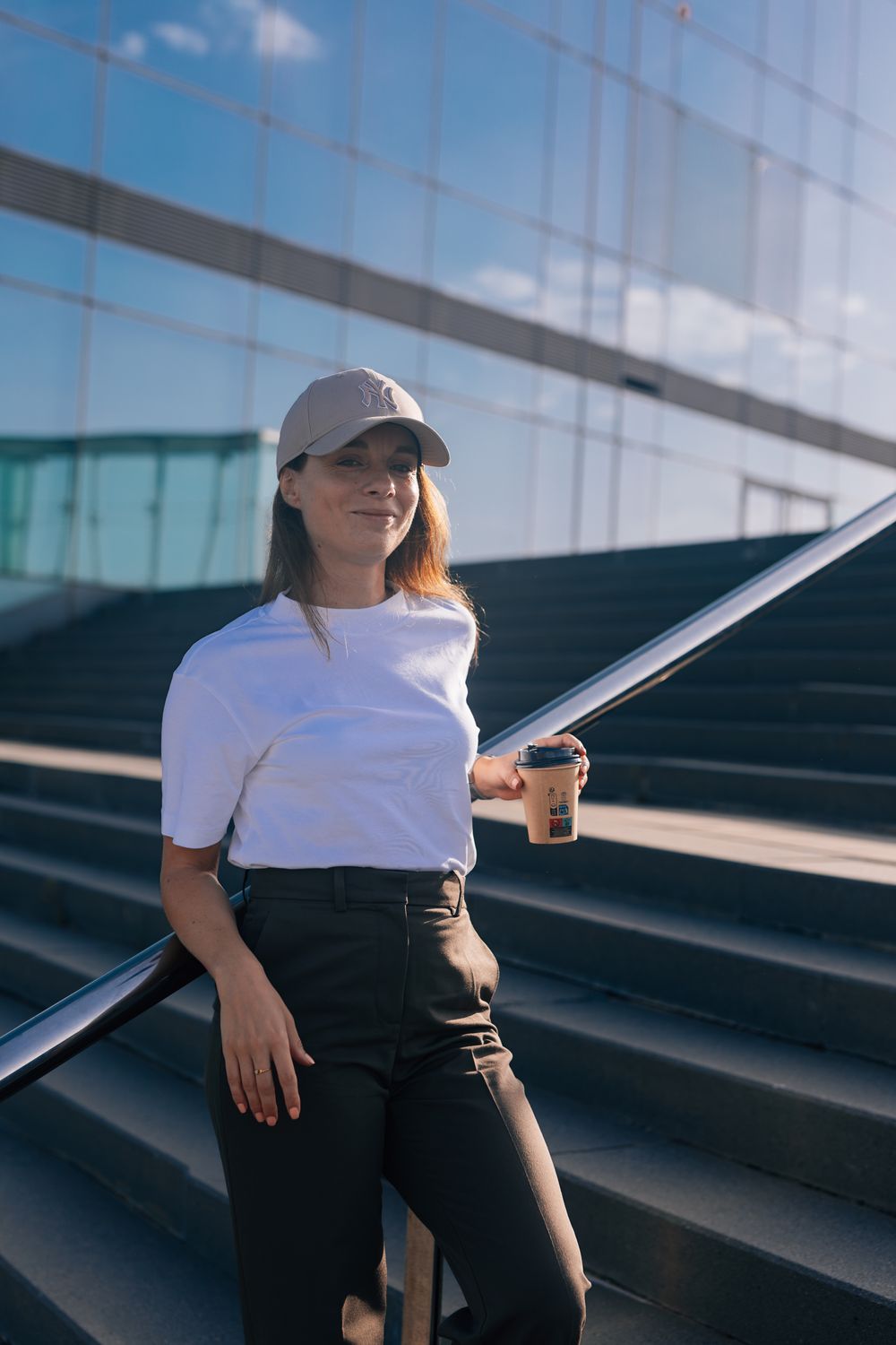 Young woman in white t-shirt and cap holding coffee cup on urban stairs with glass building backdrop.