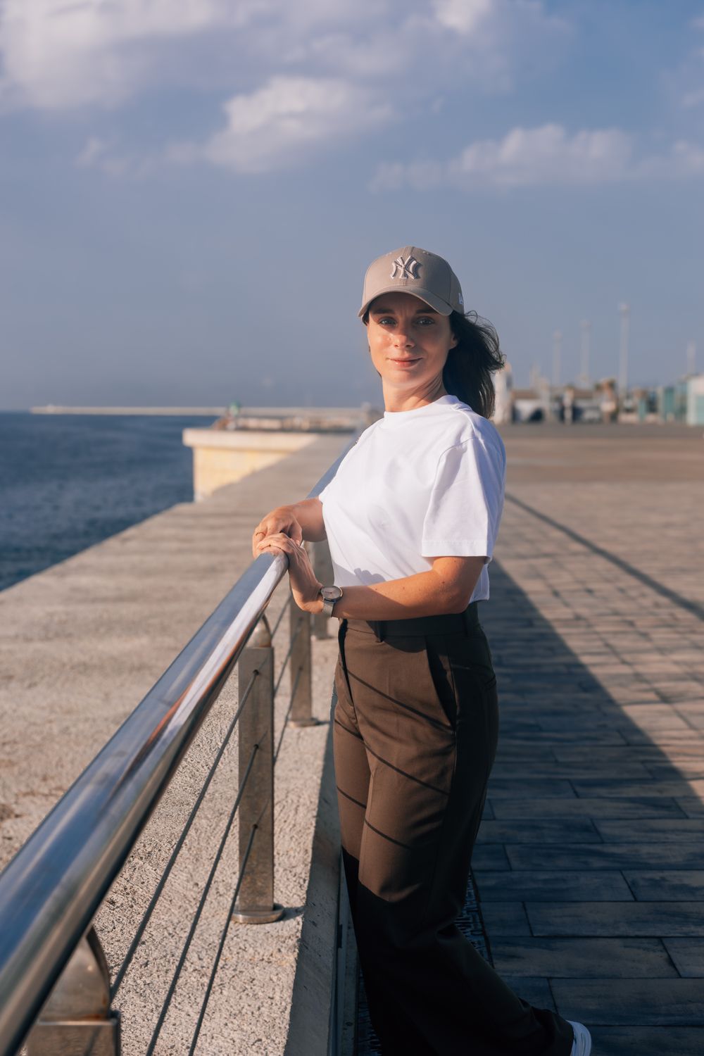 Woman in white t-shirt and NY cap leaning on railing at seaside promenade on sunny day.
