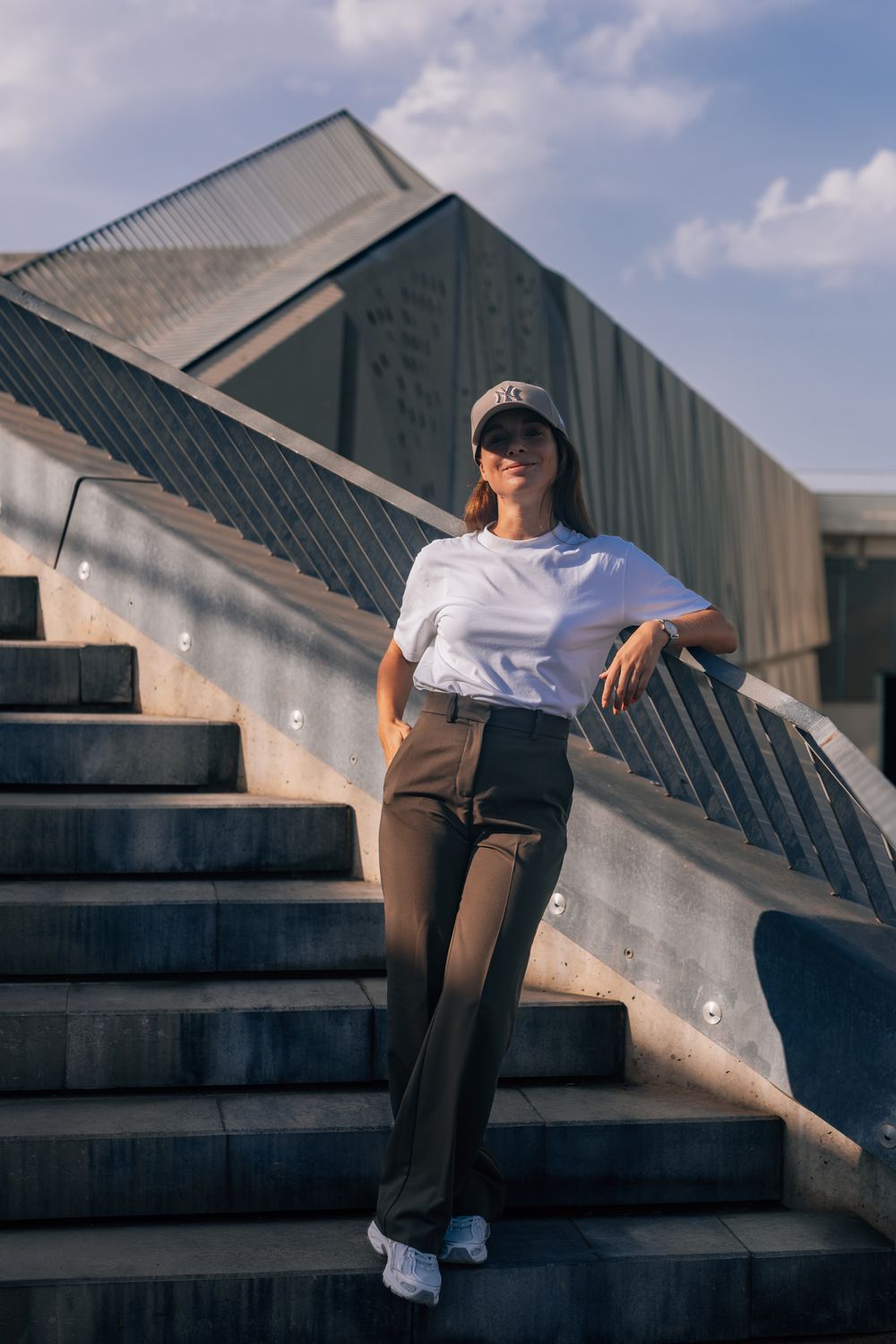 Young woman in white tee, brown trousers, and cap poses on outdoor stairs against a modern building.