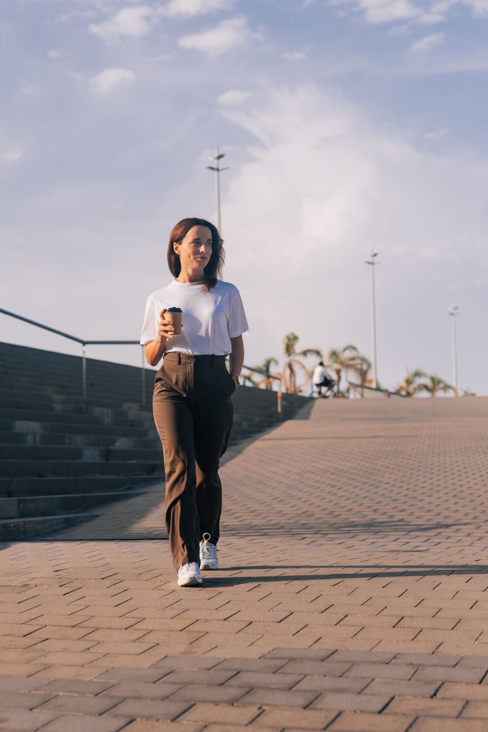 Young woman in white t-shirt and brown pants walking outdoors on a sunny day, holding a coffee cup.