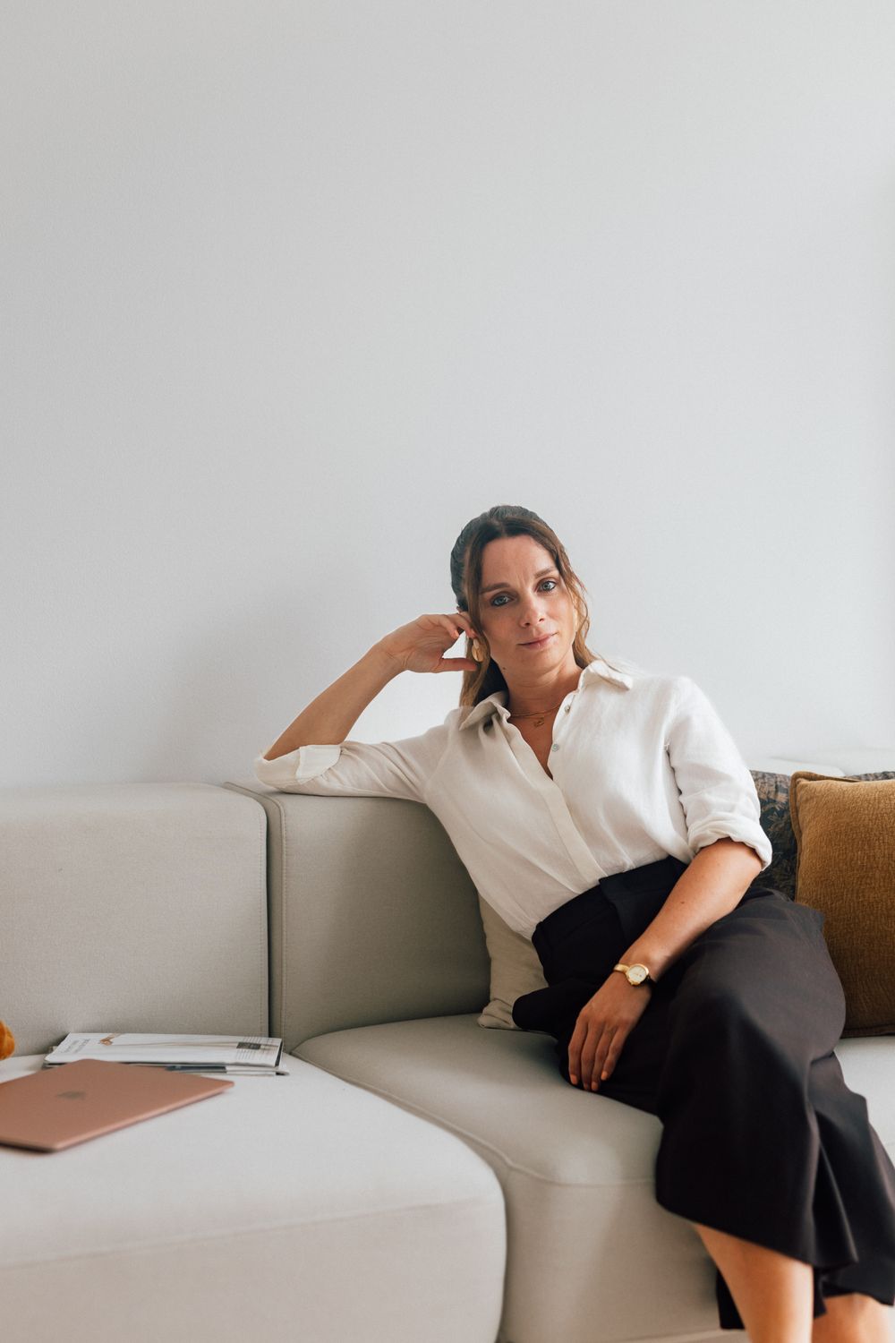 Woman in white shirt and black skirt sitting relaxed on beige sofa in minimalist living room.