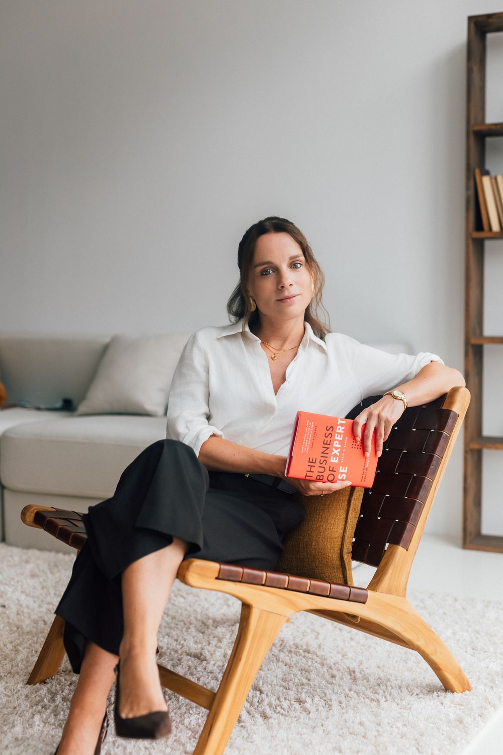 Woman in white shirt and black skirt sitting in wooden chair holding a red book in a modern living room.