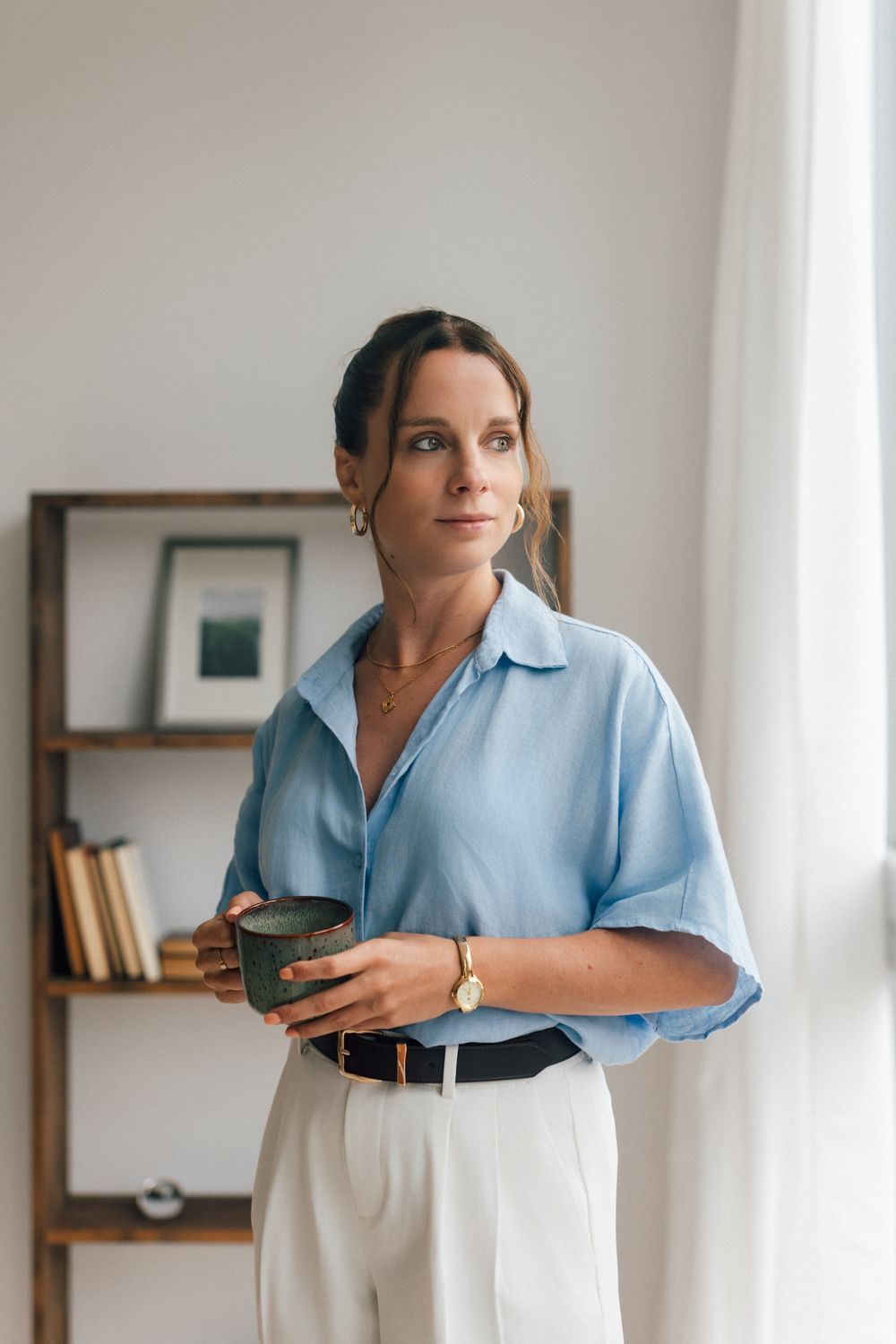 Woman in blue shirt holding a ceramic cup, standing thoughtfully near a bookshelf in a modern interior.