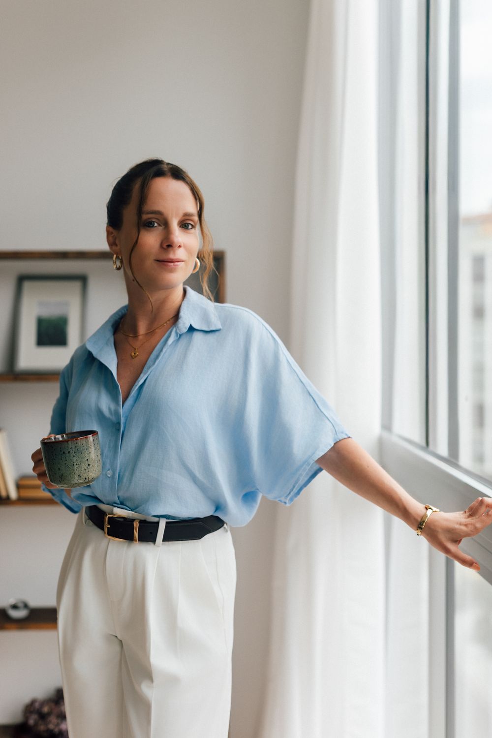 Woman in blue linen shirt and white trousers holding a mug near a bright window in a modern interior.