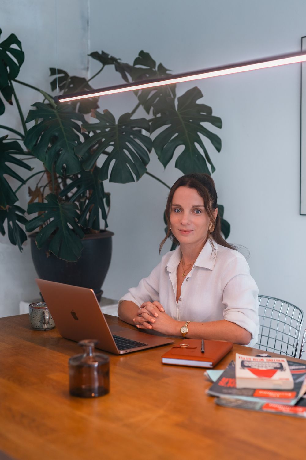 Woman in white shirt working at wooden desk with laptop, books, and monstera plant in modern office.