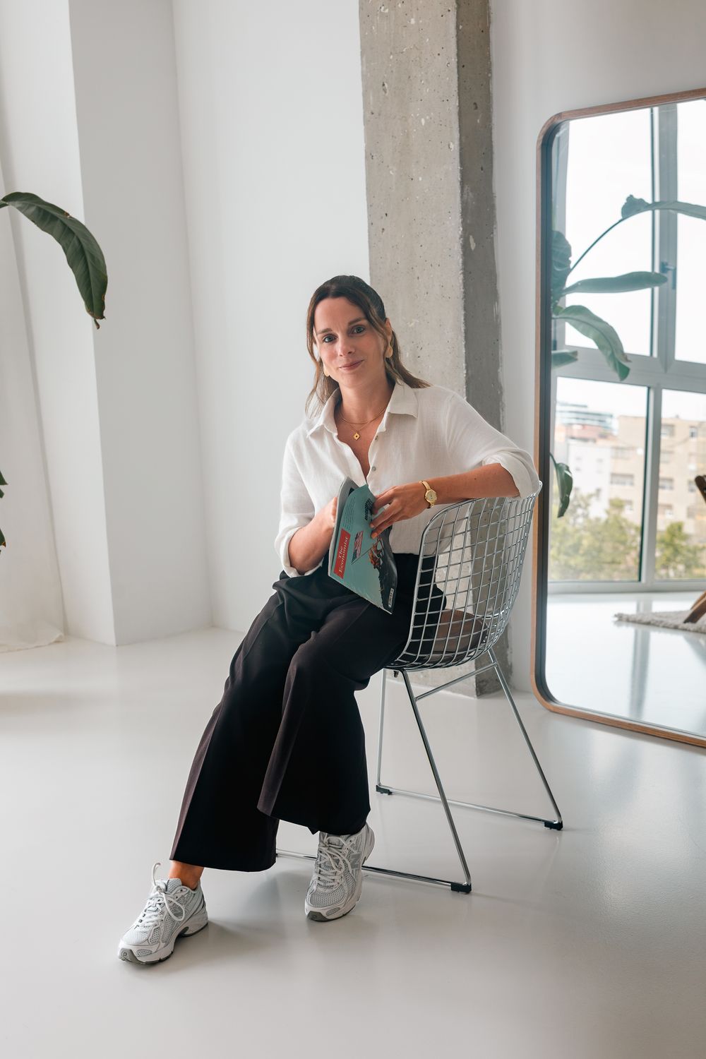 Woman in white shirt and black wide-leg pants sits in wire chair holding a book in a bright modern studio.