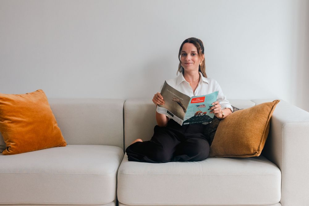 Woman sitting on a beige sofa with orange cushions, smiling and holding an open magazine in a bright living room.