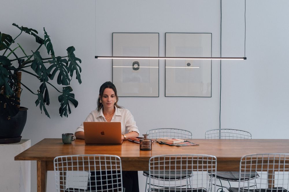 Woman working on laptop at wooden conference table in modern minimalist office with wire chairs and pendant light.