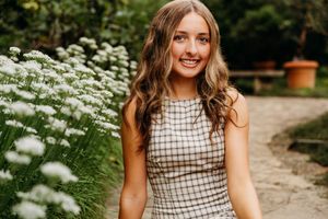 Young woman in plaid dress smiling outdoors among white wildflowers on a garden path.