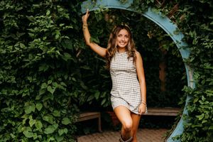 Young woman in plaid mini dress posing under a circular green ivy arch, smiling outdoors.