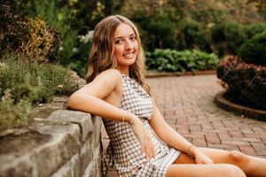 Young woman in plaid dress sitting on brick path in lush garden, smiling over shoulder.