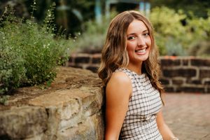 Smiling young woman in plaid dress poses by stone wall surrounded by lush greenery in outdoor garden setting.