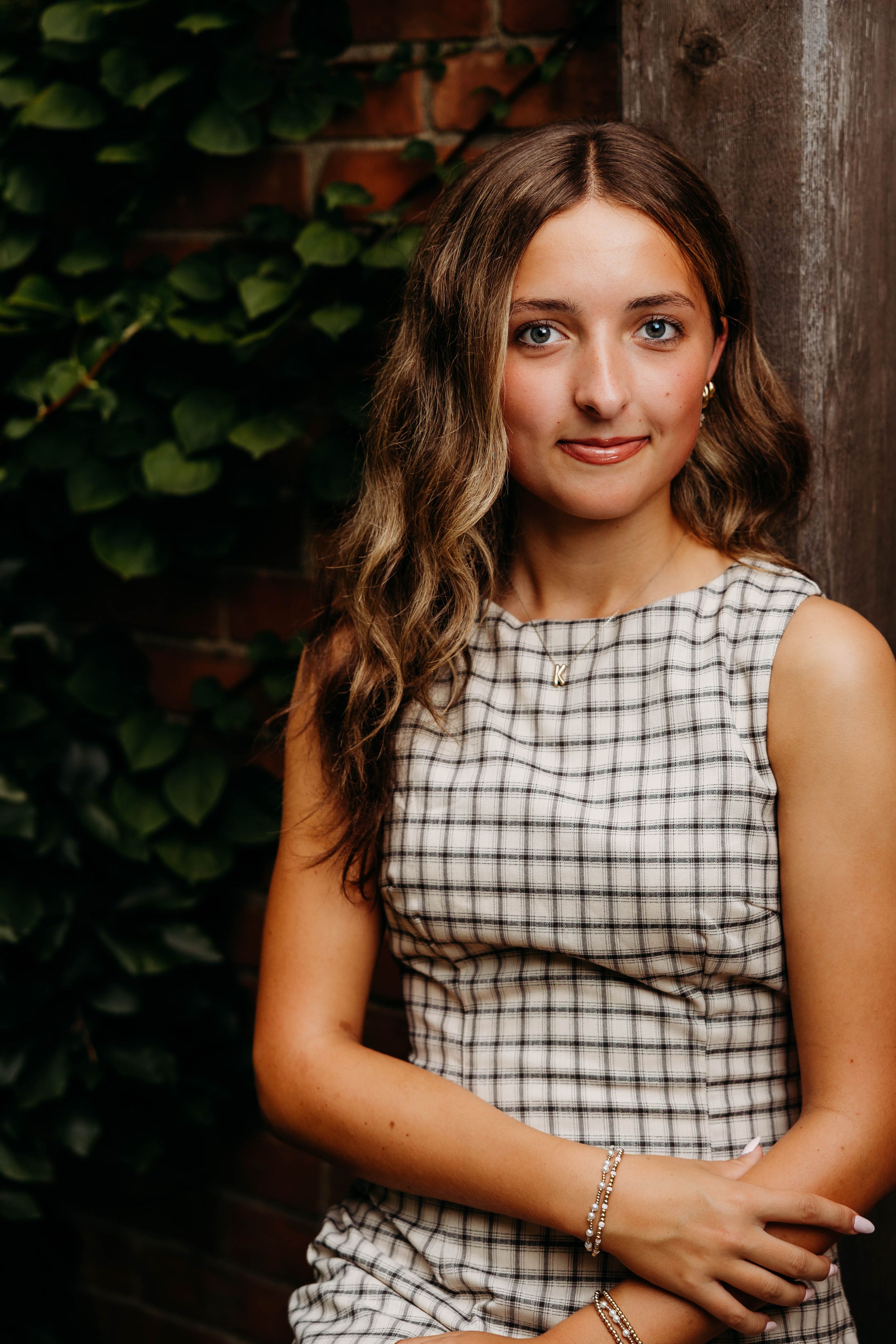 Young woman with wavy brown hair wearing a plaid dress, posing outdoors near green ivy and rustic wood wall.